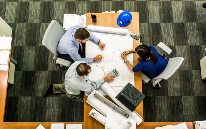 An overhead view of a meeting. Two men sit on the right of the desk and a woman in a blue top sits to the right. On the desk is a blue safety helmet, building plans, a mug, mobile phone and a laptop.